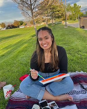 Sariah Frost sits on a picnic blanket while painting on a hand-held canvas