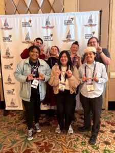 Seven students flash the U (make a U with two hands) in front of a photo backdrop that says "National LGBTQ Task Force"
