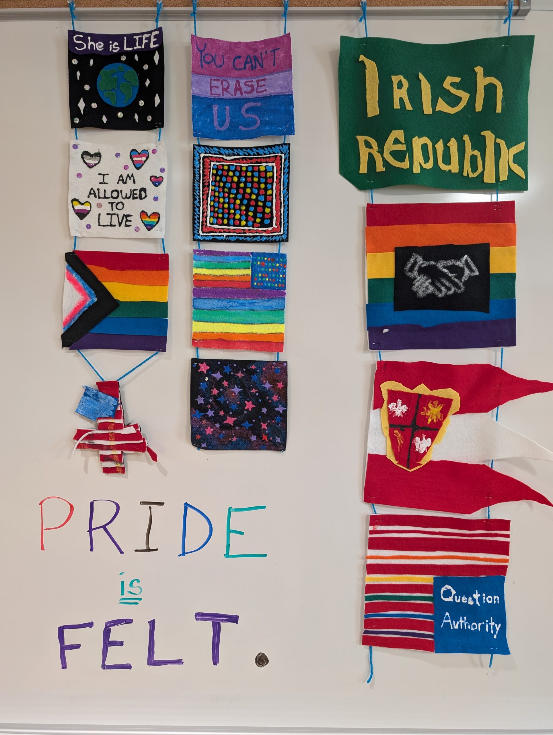 A dozen small felt shapes are hanging in front of a white board in a variety of shapes, colors, designs, and sizes to represent different aspects of student identity. Written on the white board is the sentence: "Pride is Felt."